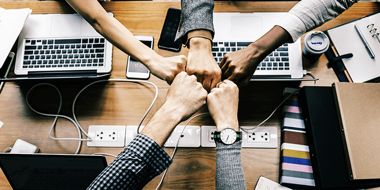 5 arms reaching in across a table in a meeting to fist bump